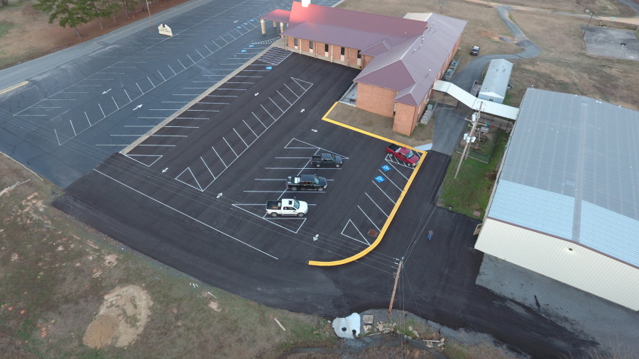 Aerial view of a mostly empty parking lot beside a large brick building, with a few cars parked near the entrance and surrounding grassy areas. A long metal-roofed structure is adjacent to the lot.