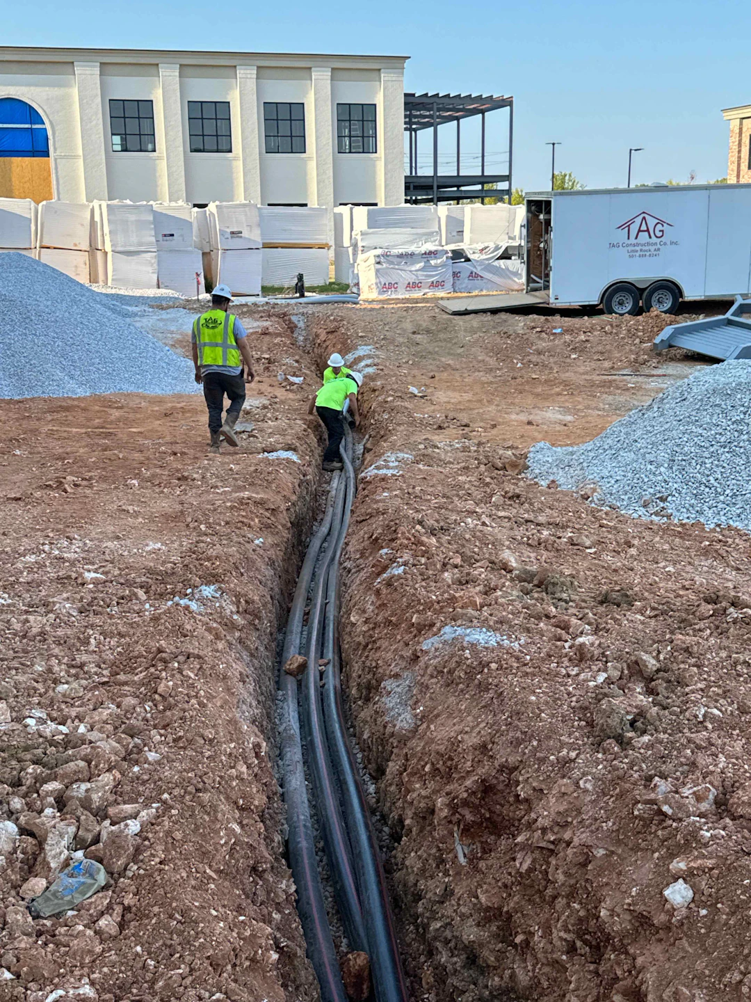 Two construction workers in safety vests and helmets work in a trench at a construction site, managing large utility cables. Piles of gravel and construction materials surround the area, with a building and trailer in the background.