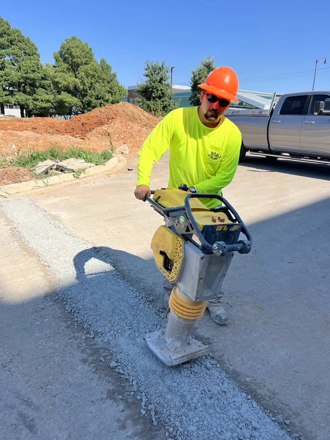 A construction worker in a bright yellow shirt and orange hard hat uses a vibrating compactor to flatten gravel on a road. Dirt piles, trees, and a parked truck are visible in the background under a clear blue sky.