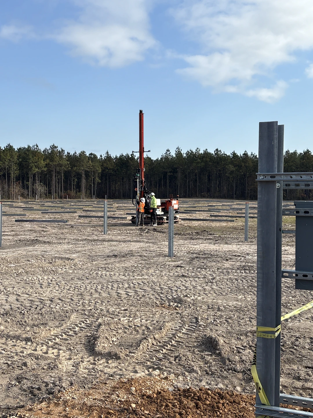 Workers operate a red drilling machine on a dirt construction site surrounded by metal posts, with a forest and blue sky in the background. Yellow tape is visible on a post in the foreground.