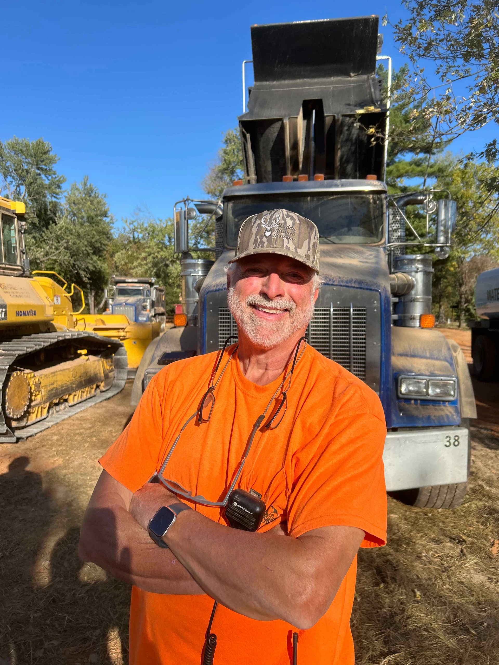 A smiling man in an orange shirt and camouflage cap stands with arms crossed in front of a large dump truck, with construction equipment and trees in the background.