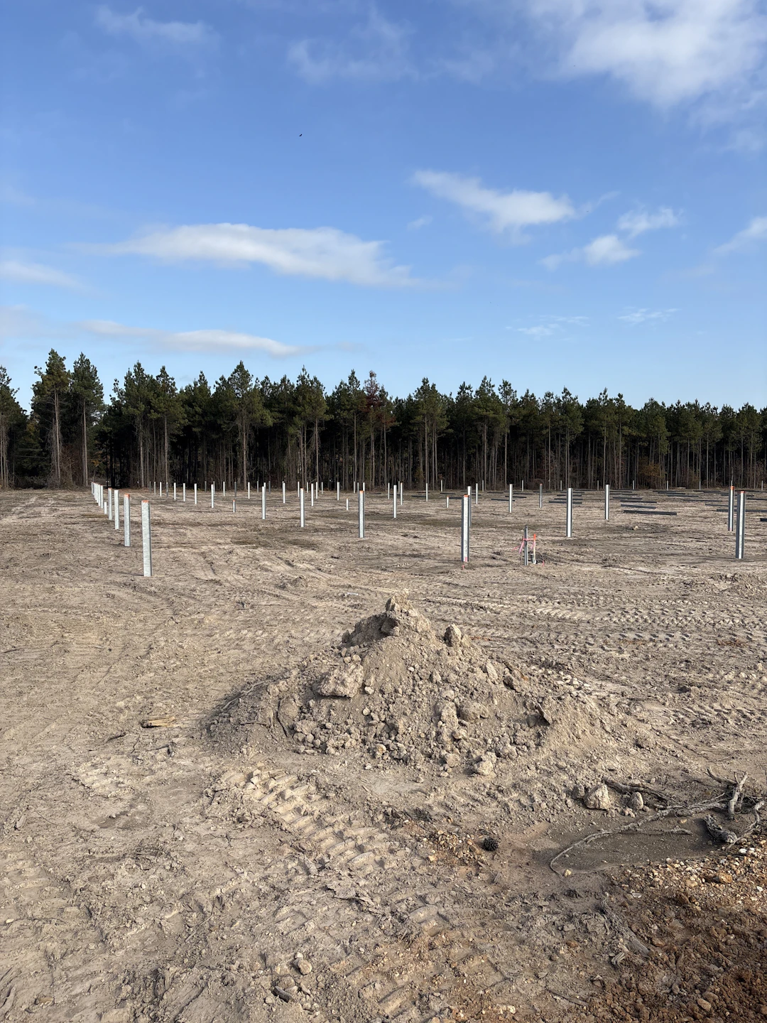 A construction site with rows of evenly spaced metal posts in the ground, piles of dirt, tire tracks, and a forest of trees in the background under a partly cloudy sky.