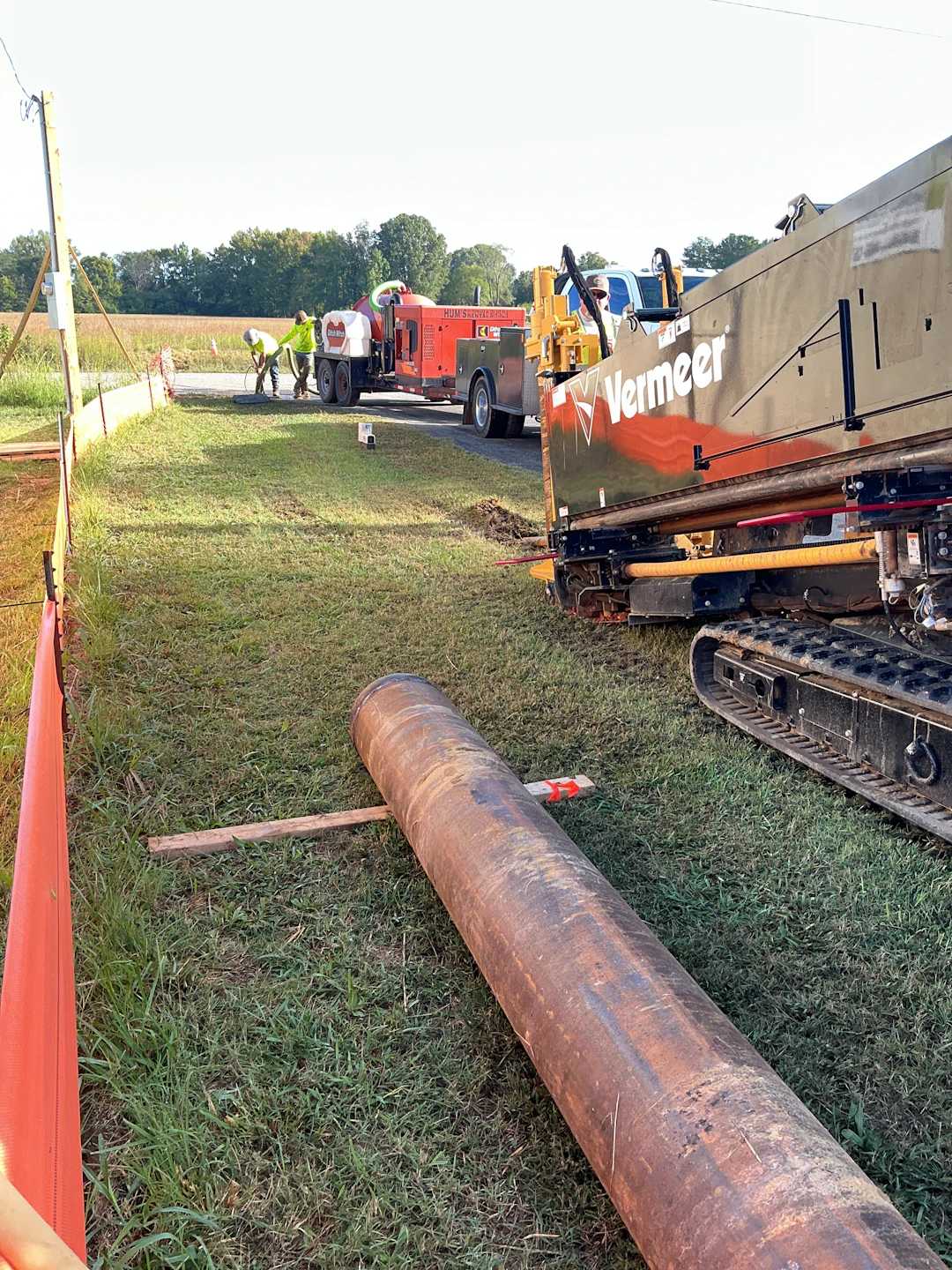 A large Vermeer construction vehicle is parked on grass near an orange safety barrier. Workers in safety vests and helmets are gathered by a red utility truck. A large metal pipe lies on the ground in the foreground.