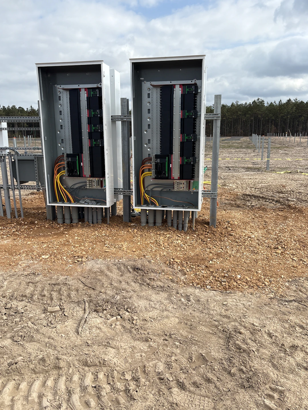 Two open electrical panels with multiple colored wires and cables are installed outdoors on bare, rocky ground near a forested area under a cloudy sky.