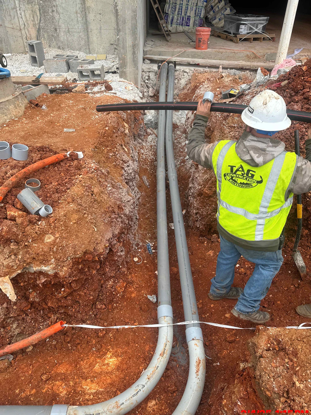 A construction worker wearing a safety vest and hard hat stands in a trench with exposed red soil, installing gray utility pipes alongside a building under construction.