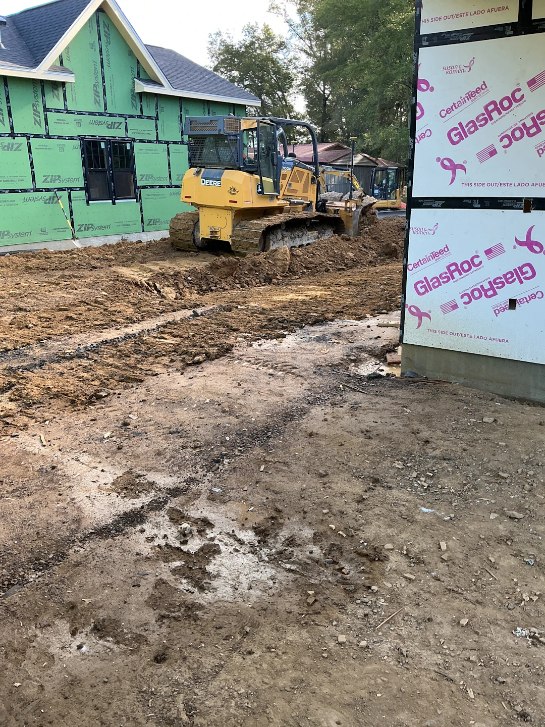 A construction worker stands beside a trench with a white pipe inside, while a small excavator operates in the background on a sunny day at a construction site with vehicles and trees visible.