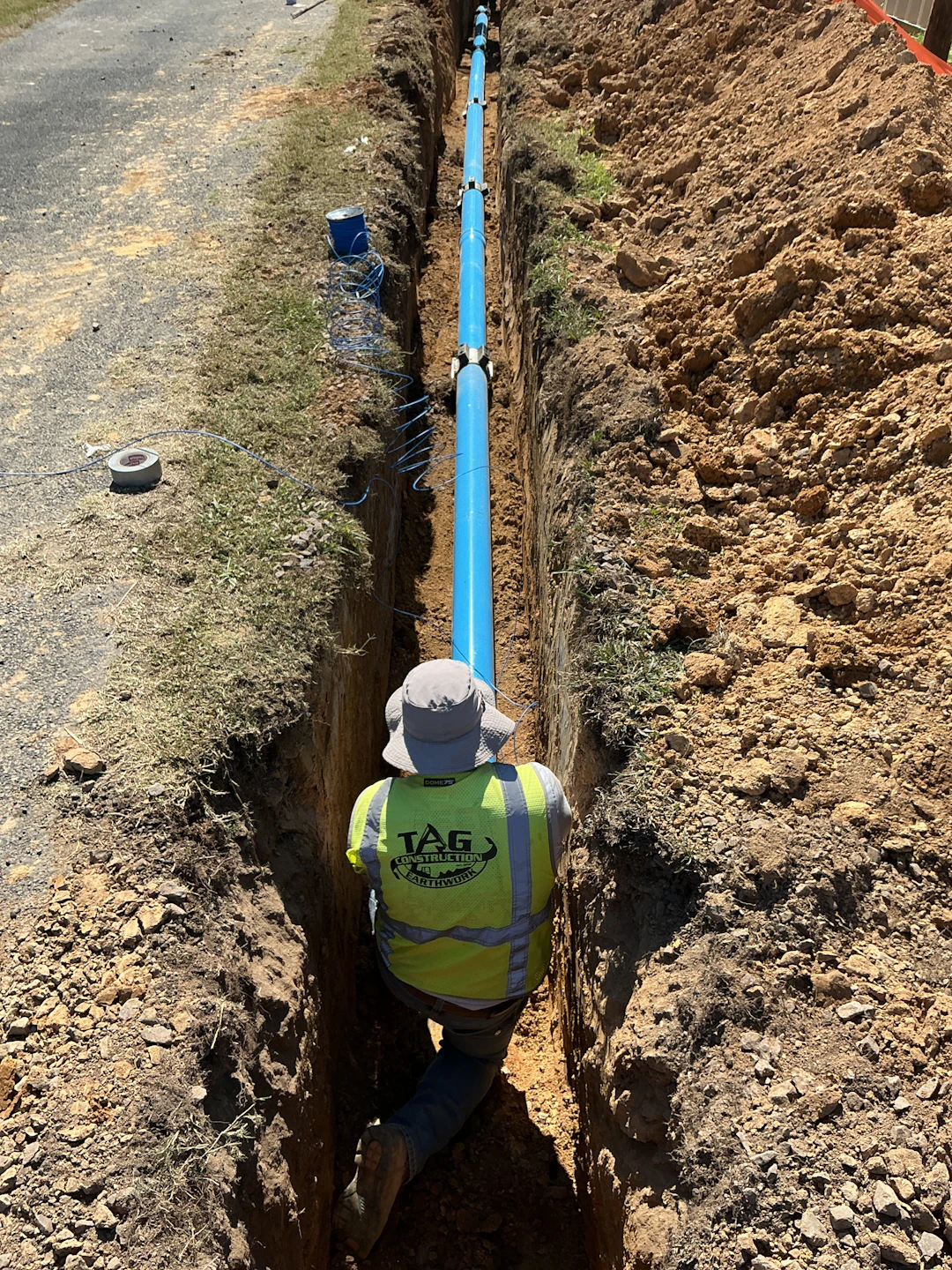A worker in a safety vest and hat kneels in a narrow trench, installing or inspecting a long blue pipe laid in the ground, with soil piled on both sides and tools nearby.