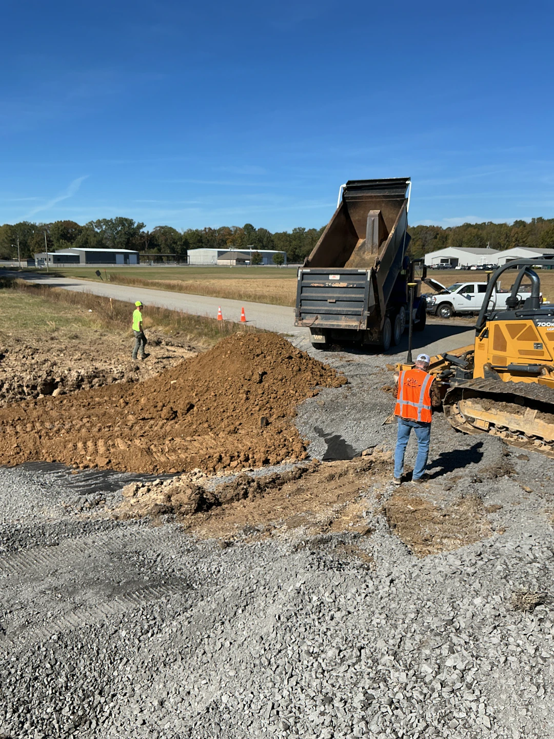 Construction workers spread dirt at a work site. A dump truck unloads soil, and a worker in an orange vest stands nearby while another operates a bulldozer. One worker in a yellow vest walks across the cleared area.