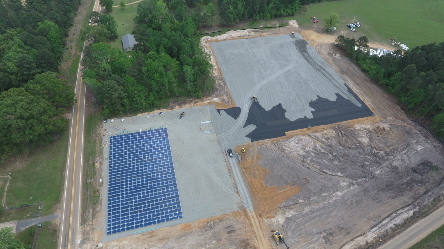Aerial view of a construction site with a large solar panel array on the left and machinery working on leveling and paving a dirt lot on the right, surrounded by trees and a nearby road.